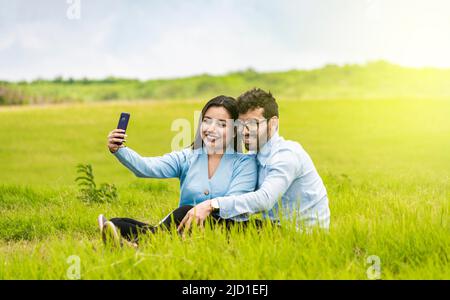 Smiling couple in love sitting on the grass taking selfies, Young couple in love taking a selfie in the field, People in love taking selfies in the Stock Photo