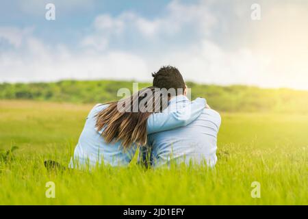 Romantic couple sitting on the grass hugging from the back, rear view of a couple in love hugging on the grass, A couple in love sitting on the grass Stock Photo