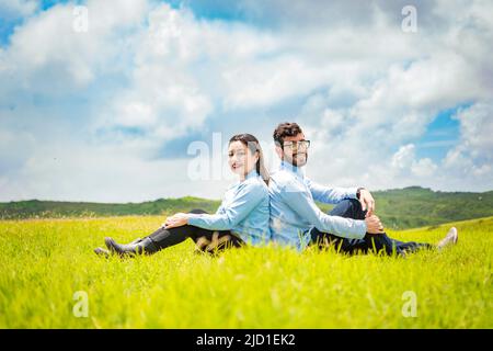 A wedding couple sitting with their backs to each other on the grass, Wedding couple in the field sitting with their backs to each other looking Stock Photo