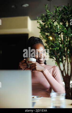 Vertical photo calm, dreaming african afro american young woman with dreadlocks, drinking coffee in cafe. Coffee break Stock Photo