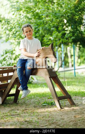 Cute little girl sitting on wooden horse in playground in green park. Summer holidays in camp, tourist center. Walking and playing outdoors, sport Stock Photo