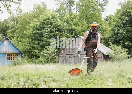 Man mowing tall grass with petrol lawn trimmer in the garden or backyard. Process of lawn trimming with hand mower Stock Photo