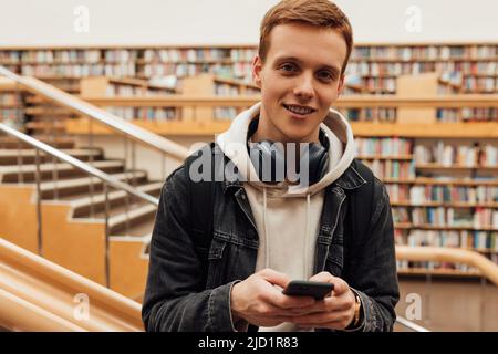 Portrait of a handsome student in casuals standing in library with smartphone Stock Photo