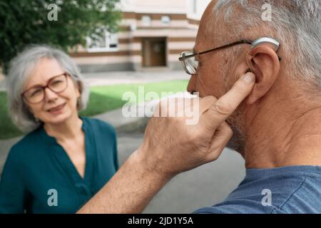 Mature man with a hearing impairment uses a hearing aid to communicate with his female senior friend outdoor. Hearing solutions Stock Photo