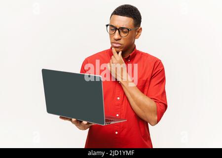 Clever and confident African-American businessman wearing smart casual shirt and eyeglasses using a laptop, a male office employee holding computer and thinking, isolated on white Stock Photo