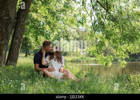 Affectionate young couple sitting on grass under tree near lake in park, looking on each other. Happy people in love Stock Photo