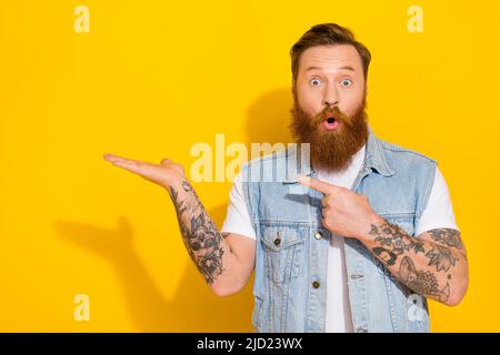 Photo of funny impressed guy dressed denim vest holding arm pointing empty space isolated yellow color background Stock Photo