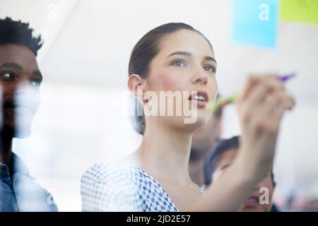 Sharing her ideas. Cropped shot of an attractive young businesswoman making notes on a glass wall while her colleagues look on. Stock Photo