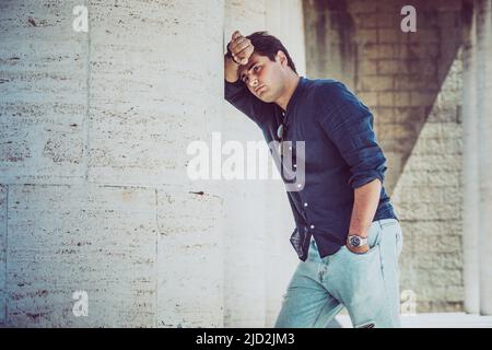 Young depressed and tired man outdoors leaning with one arm on a white wall. Casual dress. Stock Photo