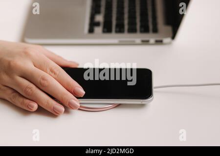 Female hand tup phone on wireless charging. Work place near at the laptop. Top view Stock Photo