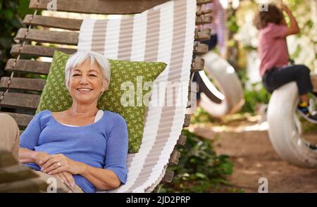 Grandmas need to recharge too. Portrait of a senior woman lying in a hammock in the garden while her grandchildren play in the background. Stock Photo