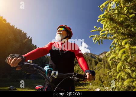 Portrait of Old cyclist with white beard holding handle bar of mountain bike in the green spruce forest in red costume in Almaty, Kazakhstan. Extreme Stock Photo