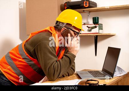 Contractor talking on cell phone while checking laptop in work office. Work organization concept, telecommunication. Stock Photo