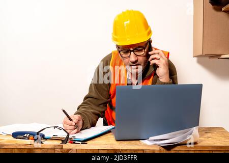 Contractor talking on cell phone while checking laptop and taking notes of what he talks about in the work office. Work organization concept, telecomm Stock Photo