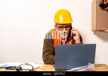 Contractor talking on cell phone while checking laptop in work office. Work organization concept, telecommunication. Stock Photo