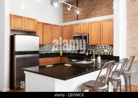 A loft kitchen with metal chairs sitting at a black granite countertop, wood cabinets, and a brick accent wall. Stock Photo