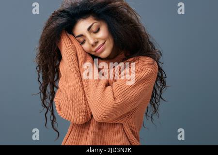 SLEEPY CONCEPT. Cheerful smiling tanned curly Latin female in warm sweater fold hands near cheek posing isolated over gray blue background close eyes Stock Photo
