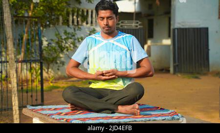 Young sporty man practicing yoga, doing Sukhasana exercise. Black pants and blue shirt, indoor full length, yoga studio Stock Photo
