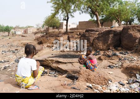 Two sad African village girls crouch on the dusty and garbage-filled roadside, among stones, sand and sparse vegetation, symbolizing the decay and des Stock Photo