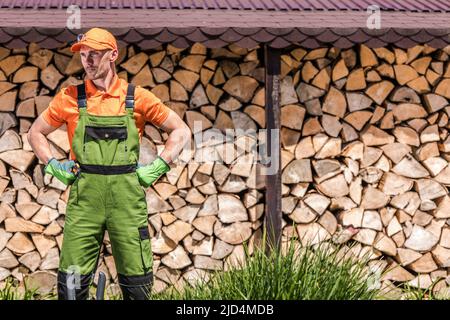 Male Caucasian Worked Dressed in Professional Gardening Wear Standing in Front of Firewood Wall Made of Chopped Wood Pieces. Stock Photo