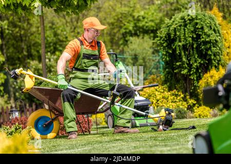 Caucasian Landscape Gardener Resting While Mowing the Lawn Sitting on the Wheelbarrow and Holding Professional Grass Trimmer in His Hands. Stock Photo