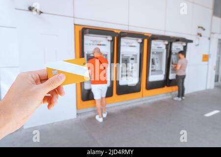 Hand holds a bank plastic card with a chip against the background of a number of ATMs in which customers withdraw or deposit money into the account Stock Photo