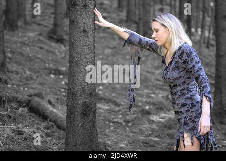 Stylish blond woman in blue designer dress posing in woodland resting her hand on the trunk of a tree in a fashion portrait Stock Photo