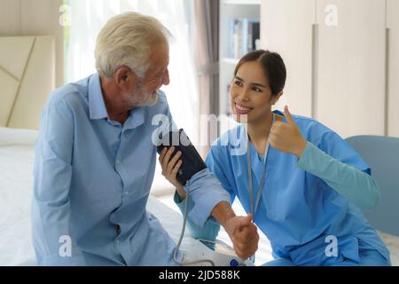 Asian Attentive caregiver using tonometer while elder man having breath problem. Female doctor measuring blood pressure of senior man at home. Stock Photo