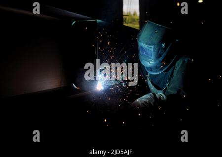 A welder in protective overalls and a welding mask welds metal structures by welding on a semi-automatic line, blue sparks fly to the sides Stock Photo