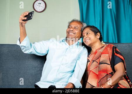 happy senior couple taking selfie on mobile phone while sitting on sofa at home - concept of family, togetherness and bonding. Stock Photo