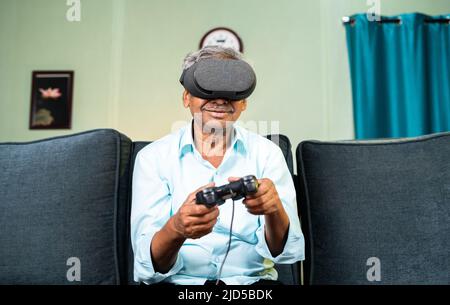 happy smiling Old man experiencing VR headset by playing video game using gamepad at home while sitting on sofa - concept of technology and Stock Photo
