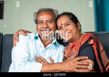 Happy smiling senior Indian couple embracing each other by looking at camera while on sofa - concept of family bonding, relaxation and happiness Stock Photo