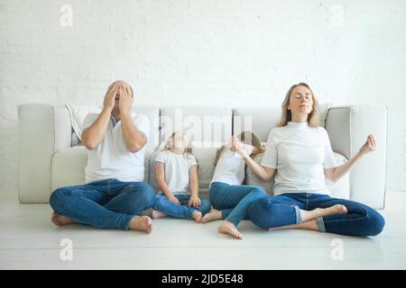 Calm mother with closed eyes meditating in lotus pose on floor, stressed father and two tired children in living room at home. Stock Photo