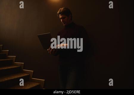 Confident portrait of a serious handsome mature Caucasian businessman, entrepreneur, copywriter working online on laptop standing against a dark wall Stock Photo