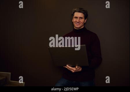 Confident portrait of a serious handsome mature Caucasian businessman, entrepreneur, copywriter working online on laptop standing against a dark wall Stock Photo