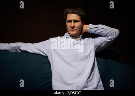 Relaxed handsome attractive Caucasian man in casual wear looking at camera, sitting on a new comfortable couch, testing it while shopping for new sofa Stock Photo