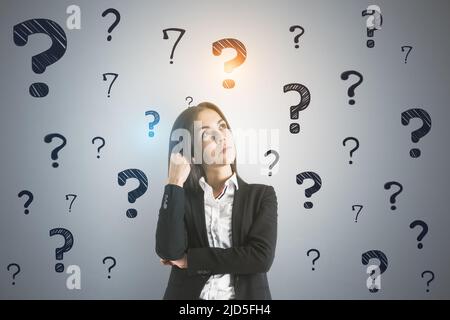 Searching for an answer concept with pensive young businesswoman on light wall background with black question marks Stock Photo