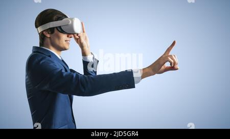 Young businessman working in virtual reality office in VR glasses, presses his finger virtual interface on abstract light blue backdrop. Innovate and Stock Photo