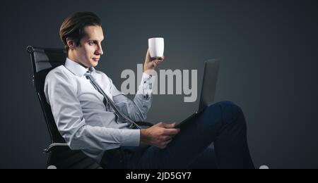 Distant work and freelance concept with man in white shirt sitting on office chair with cup of coffee in hand and looking at laptop screen on dark gre Stock Photo