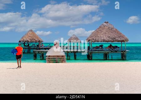 A young man walking down a beautiful beach with  people standing on a wooden jetty on Cayo Guillermo, Cuba Stock Photo