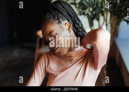 Young afro american woman stretching neck with closed eyes tilting head to side, sitting and working in coffee house. Black girl working remotely Stock Photo