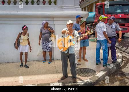 An old street musician plays guitar on the pavement and is surrounded by multiple people in Santiago de Cuba, Cuba Stock Photo