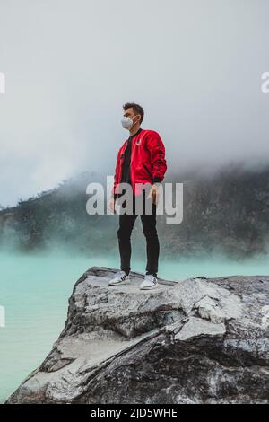 young man in red bomber jacket standing on rock at kawah putih sulfer lake Stock Photo