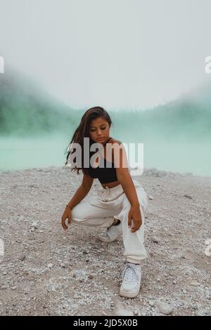 young Indonesian woman kneeling at sulfur lake Kawah Putih surrounded by green fog Stock Photo