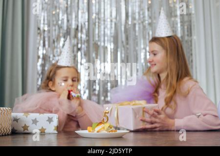 Two sisters celebrate birthday of youngest girl of 5 years. Girls are dressed in party hats and dresses, older one holds gift box in her hands