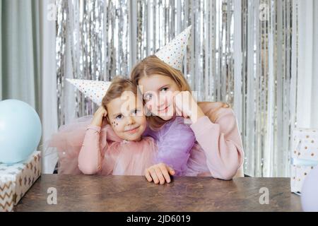 Portrait of two charming sisters celebrating their birthday. Girls in party hats and festive dresses sit same way, with their cheeks propped up with