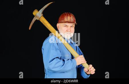 Bearded man in uniform with pickaxe. Male builder in hard hat with old pick mattock. Mining device. Stock Photo