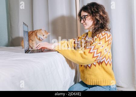 A stylish female freelancer in a yellow sweater and glasses works at home on a laptop sitting on the floor with her pet red cat. Remote work from home during lockdown. Stock Photo
