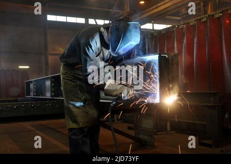 A welder in protective overalls and a welding mask welds metal structures by welding on a semi-automatic line, blue sparks fly to the sides Stock Photo