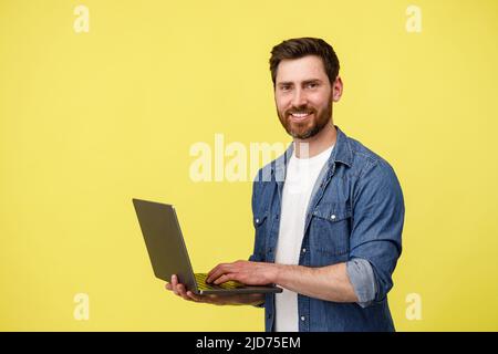 Handsome smiling man with beard holding laptop and looking at camera on yellow background Stock Photo